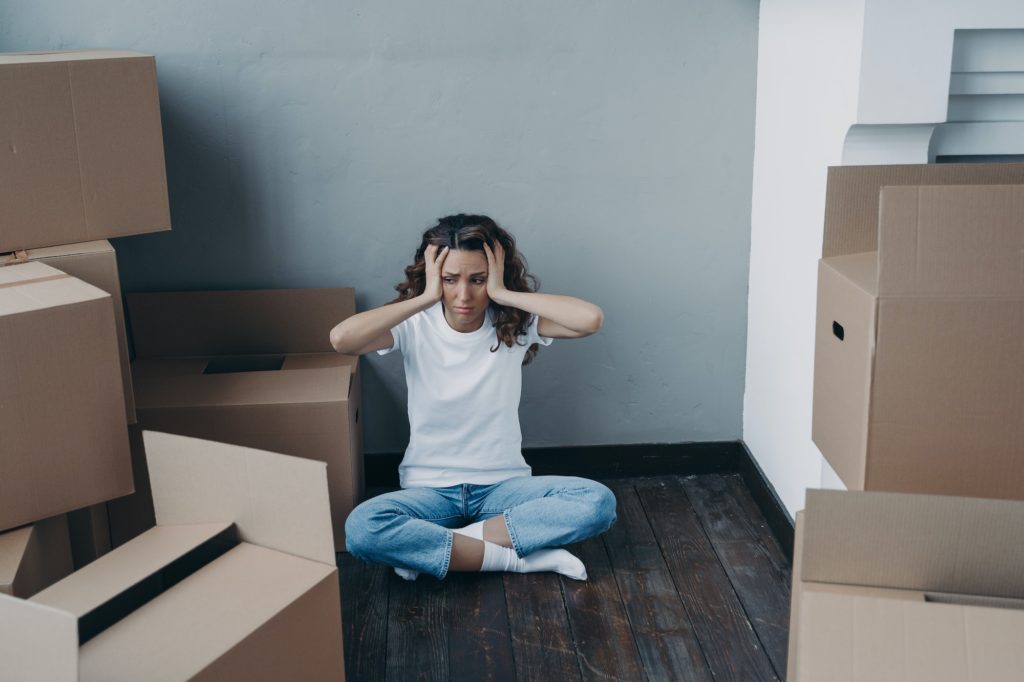 Exhausted young woman amond packed boxes. Stress, headache and depression from moving.